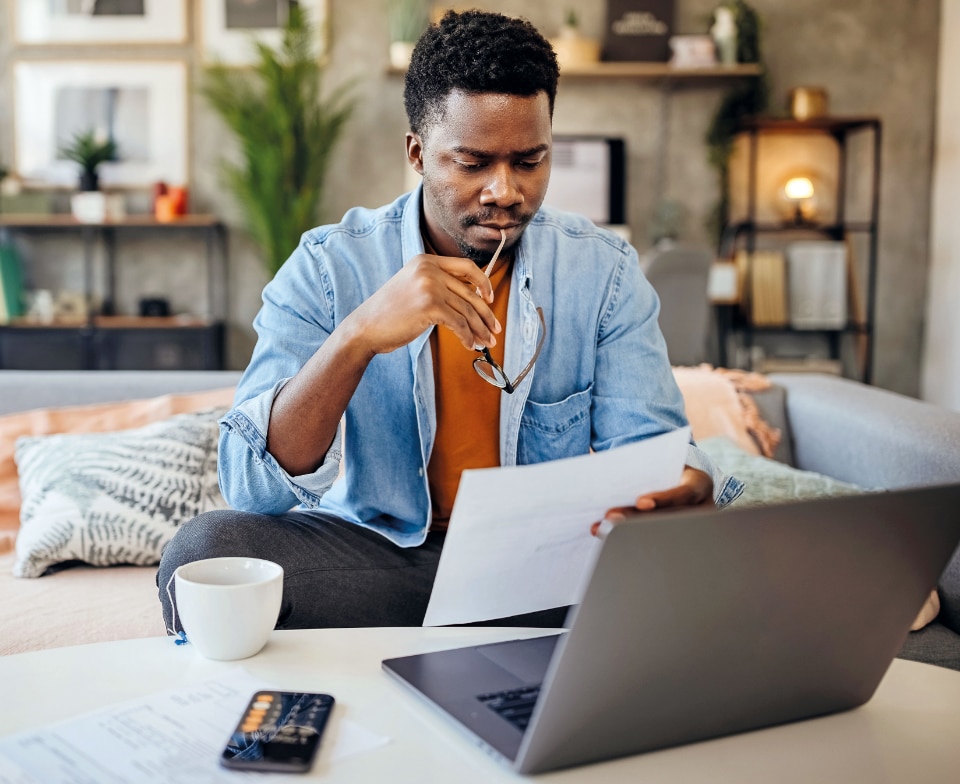A foto mostra a sala de uma casa onde vemos um jovem negro usando camiseta laranja e camisa jeans por cima. Ele encara a tela do notebook enquanto segura uma conta em uma das mãos e os óculos na outra.