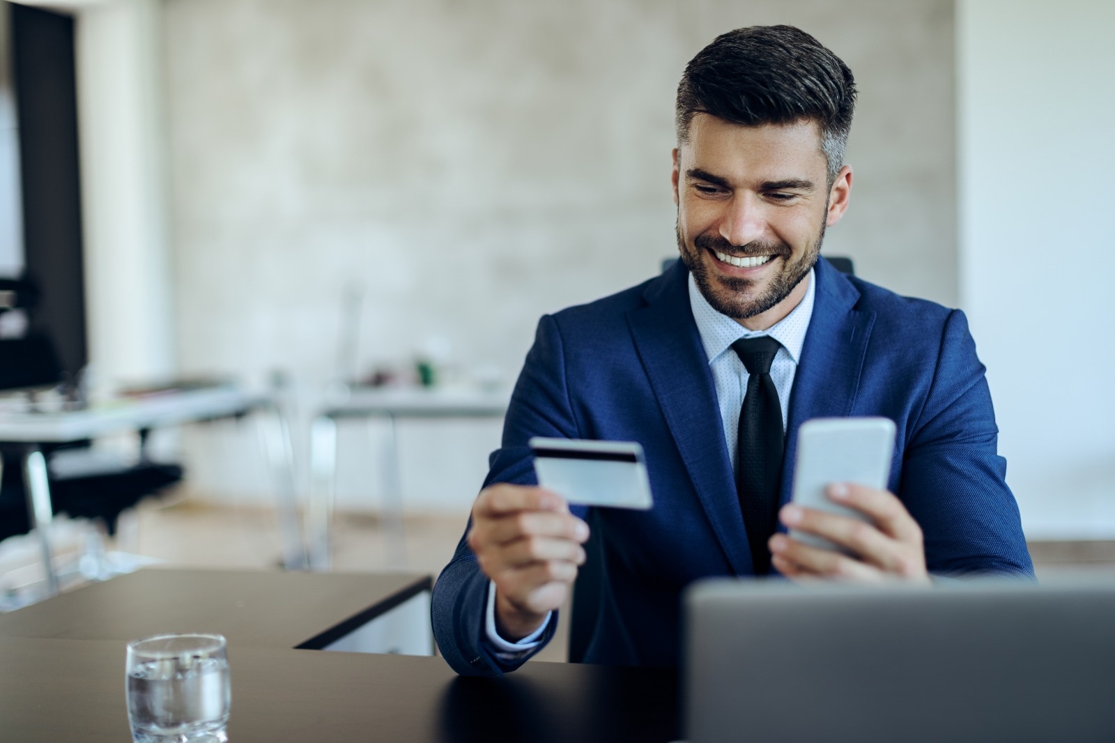 Pessoa sentada em uma mesa de escritório, usando terno azul, segurando um cartão bancário em uma mão e um smartphone na outra. Há um notebook desfocado em primeiro plano e um copo de água sobre a mesa. Ao fundo, o ambiente corporativo aparece claro e desfocado.