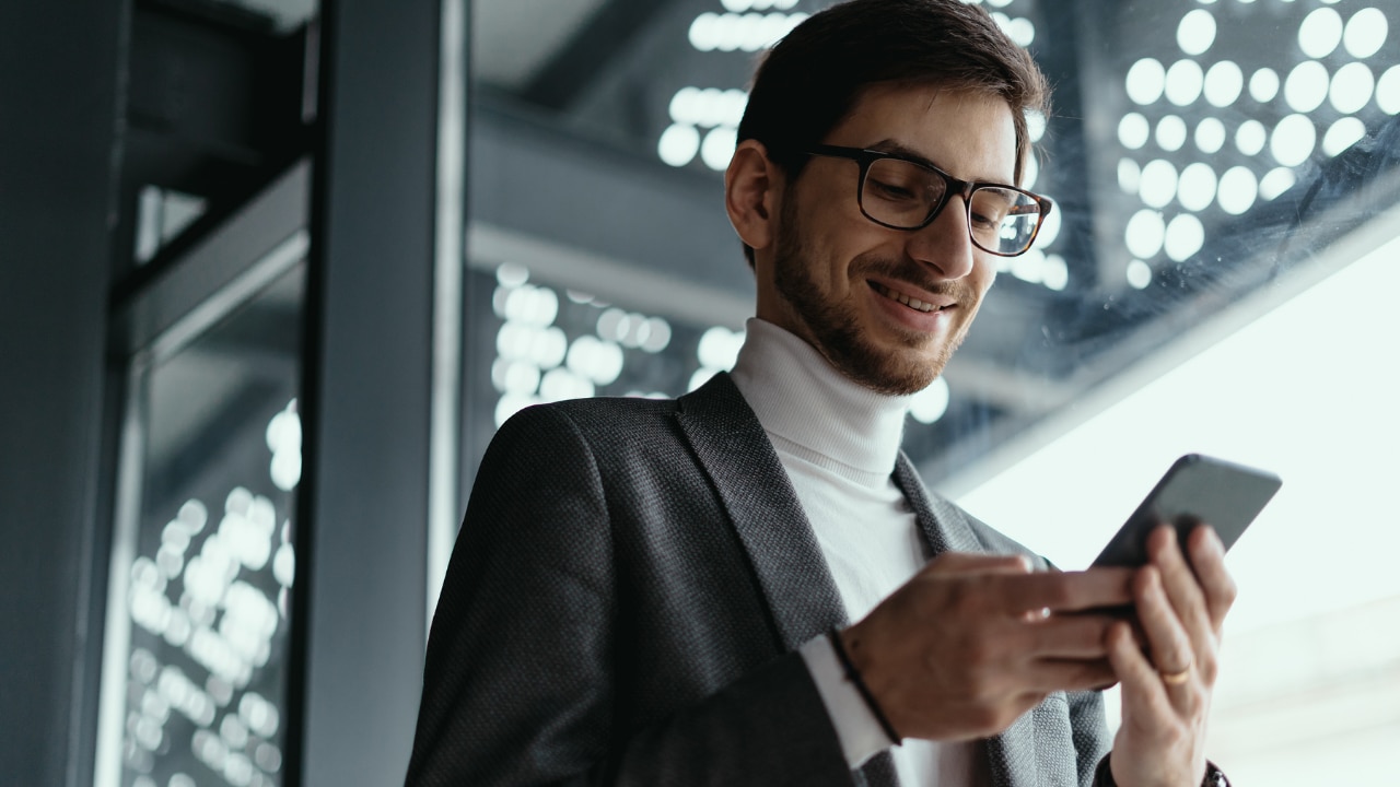 Pessoa em ambiente corporativo segurando um smartphone e interagindo com a tela, em um espaço interno moderno com estrutura de vidro e iluminação natural ao fundo.