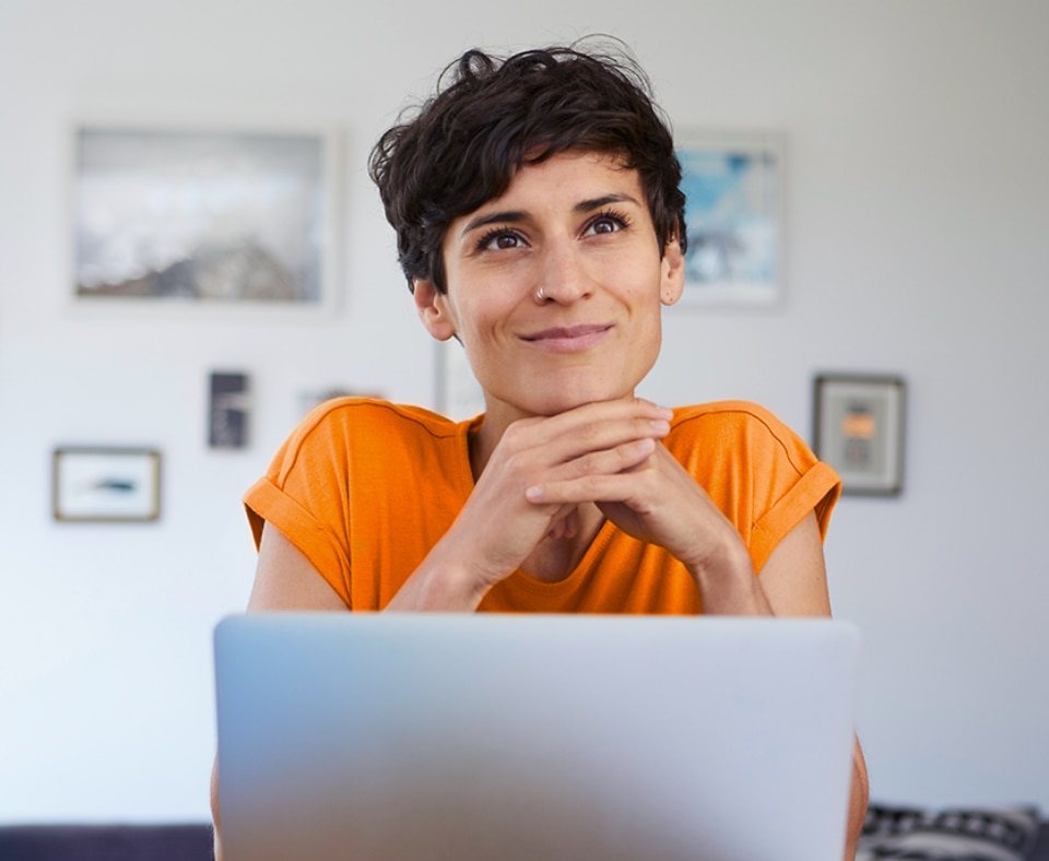 #ParaTodosVerem: Uma moça branca, de cabelo curto preto está com o laptop aberto na mesa de casa. Ela sorri levemente.