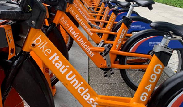 Fila de bicicletas laranjas do Bike Itaú Uniclass estacionadas lado a lado em área urbana.